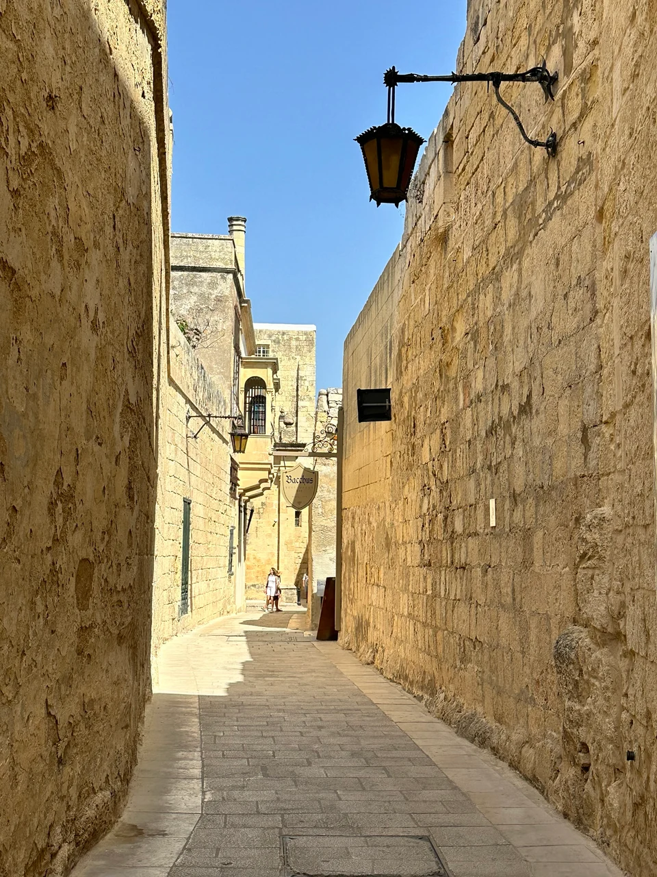 A narrow sunlit limestone alley with a wrought-iron lantern on the wall and a person walking in the distance.