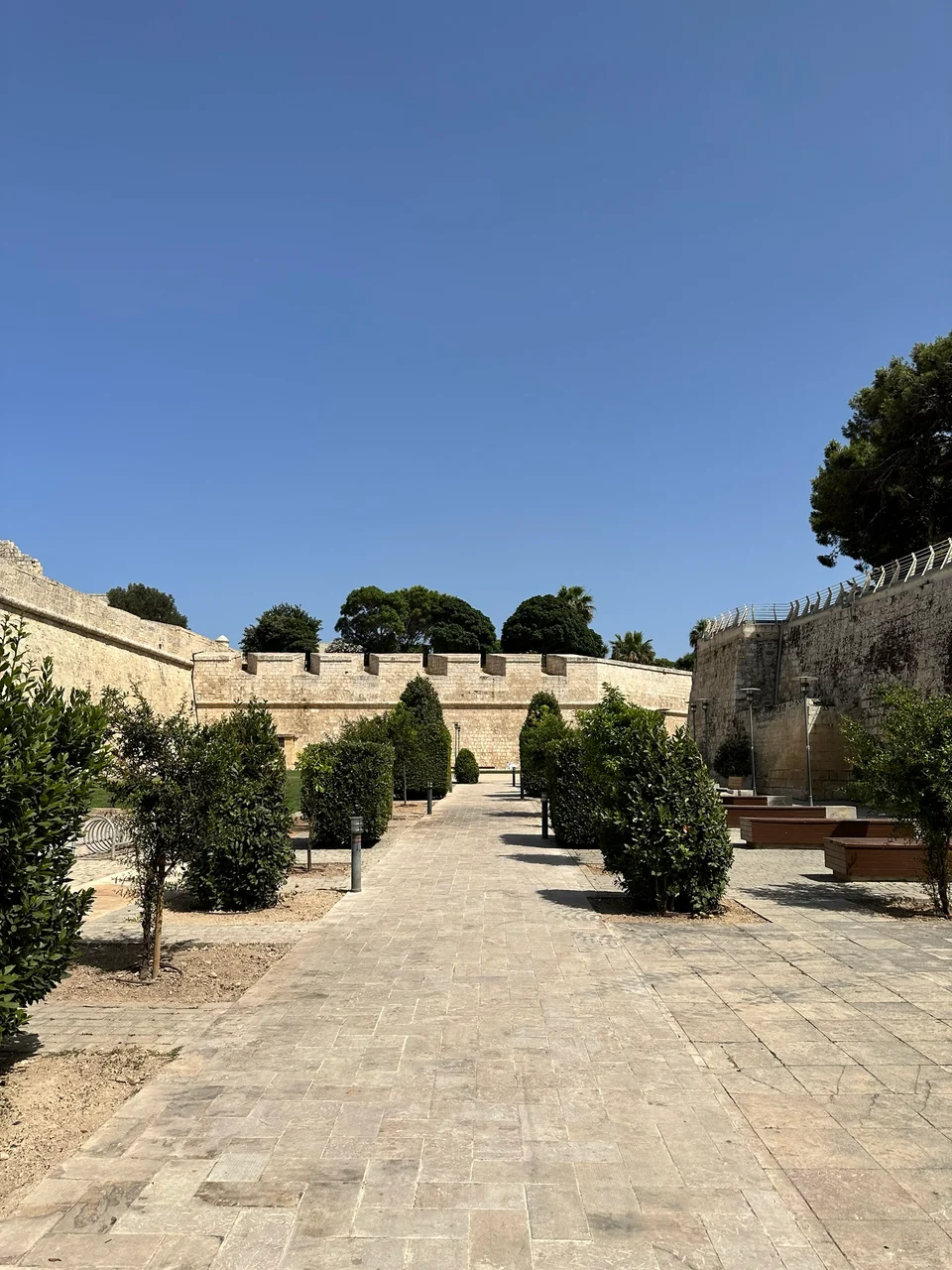 A stone-paved path lined with trimmed shrubs leads towards ancient limestone battlements under a clear blue sky.