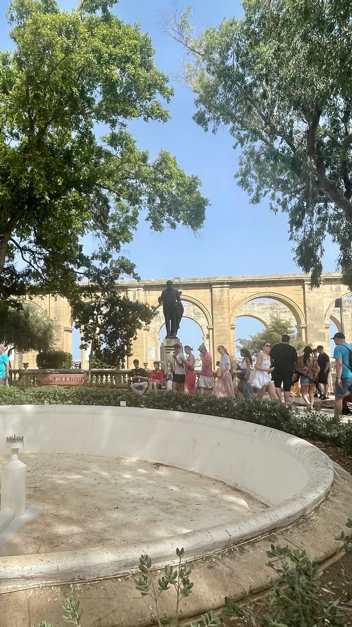 Tourists walk past a bronze statue and stone arches in a sunny garden with a dry fountain in the foreground.