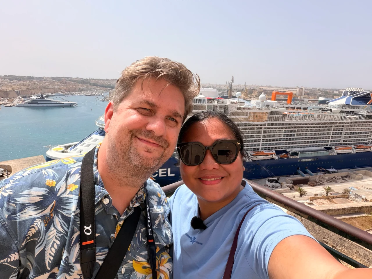Two people take a selfie from a high viewpoint overlooking a harbour with large cruise ships docked below.