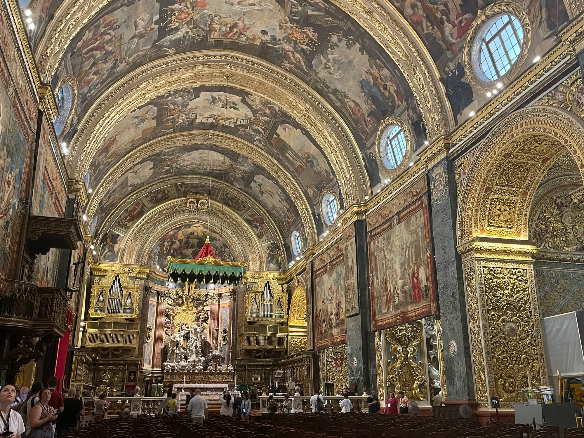 The ornate gilded interior of a baroque cathedral with a vaulted ceiling covered in painted frescoes and rows of dark wooden pews.