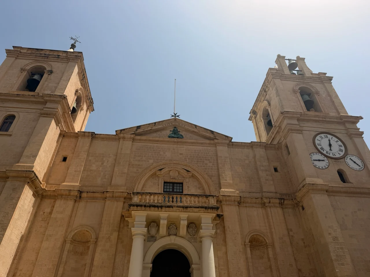 Looking up at the pale stone facade of St John's Co-Cathedral with two bell towers and a clock face against a blue sky.
