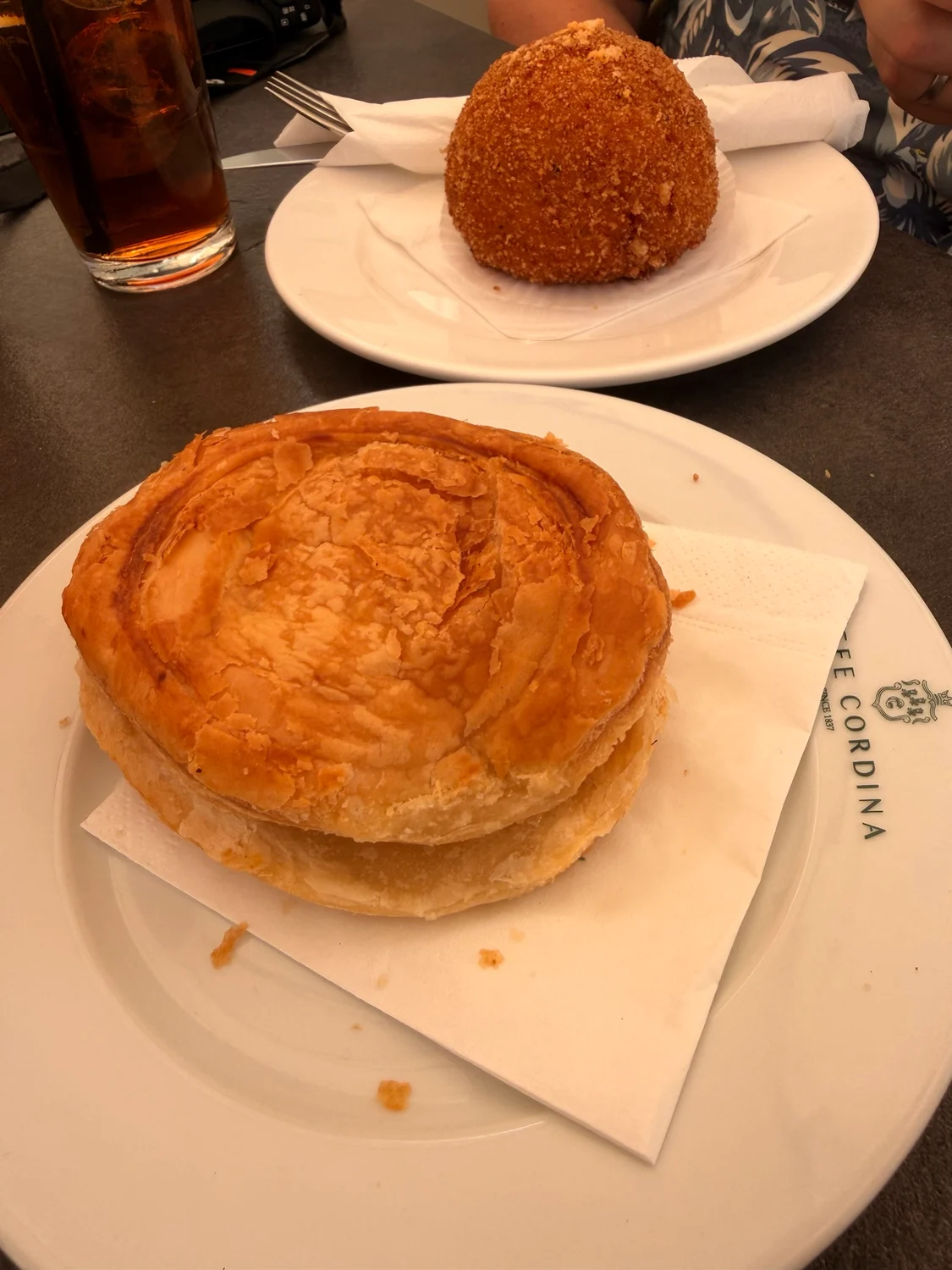 Two white plates on a dark table, one holding a large flaky pastry pie and one a round breaded ball, with a napkin marked Cafe Cordina.