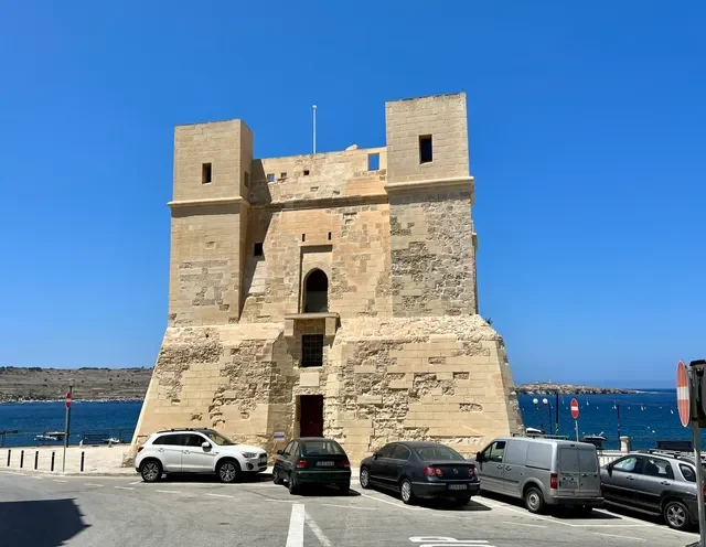 A large square limestone coastal tower with two corner turrets, cars parked in front and blue sea behind.