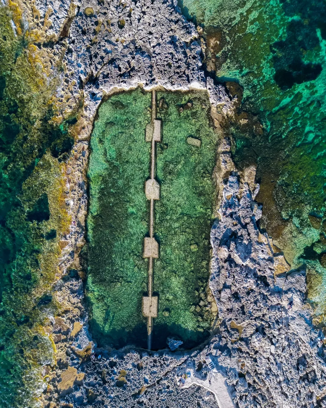Aerial view of a rectangular rock pool filled with clear green water, with a narrow walkway running down its centre.