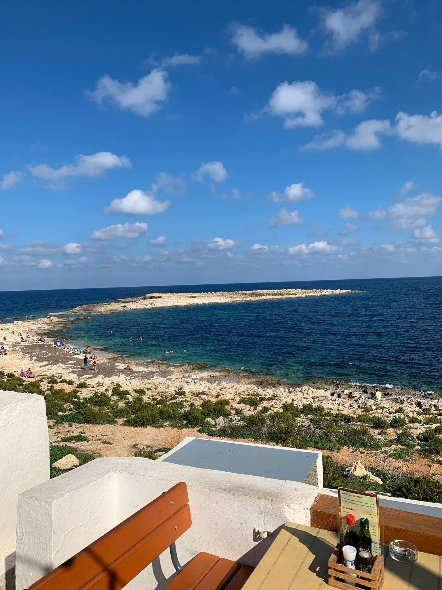 A terrace table overlooks a rocky Mediterranean cove with turquoise water, a limestone headland and a blue sky with scattered clouds.