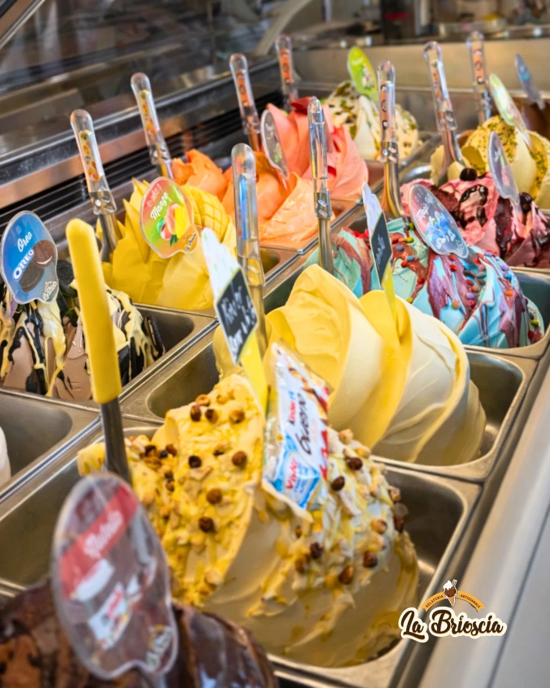 A gelato display counter showing tubs of colourful ice cream in flavours including Oreo and mango, each with a spade and label.