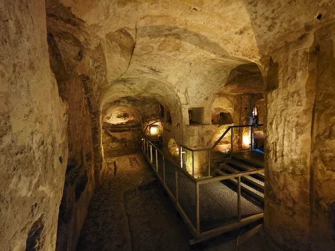 A dimly lit ancient stone tunnel with vaulted ceilings, modern metal walkways and steps, and warm amber lighting.