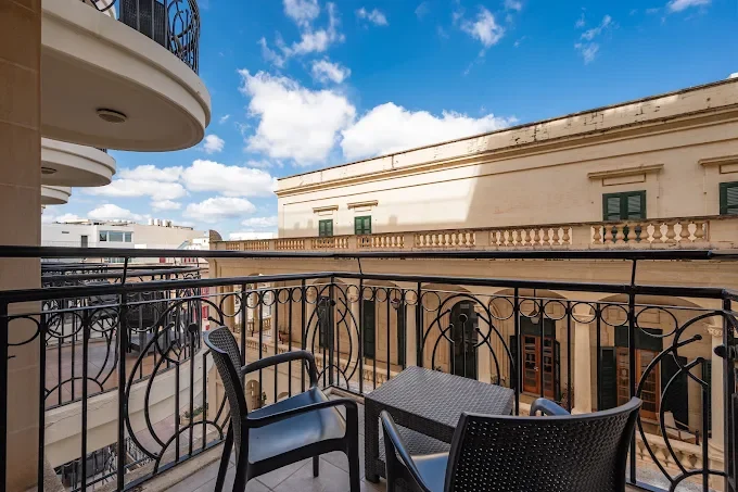 A wrought iron balcony with two wicker chairs and a small table overlooks a street lined with classical stone buildings.