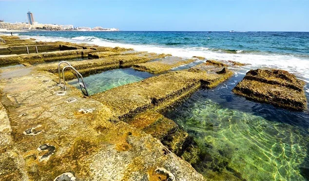 Rocky coastal pools carved into limestone at the sea's edge, with clear green water and a metal handrail, and a town in the distance.