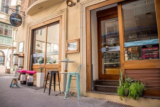 Exterior of Kapitali bar in Valletta with bar stools on the pavement and a wooden-framed entrance door.