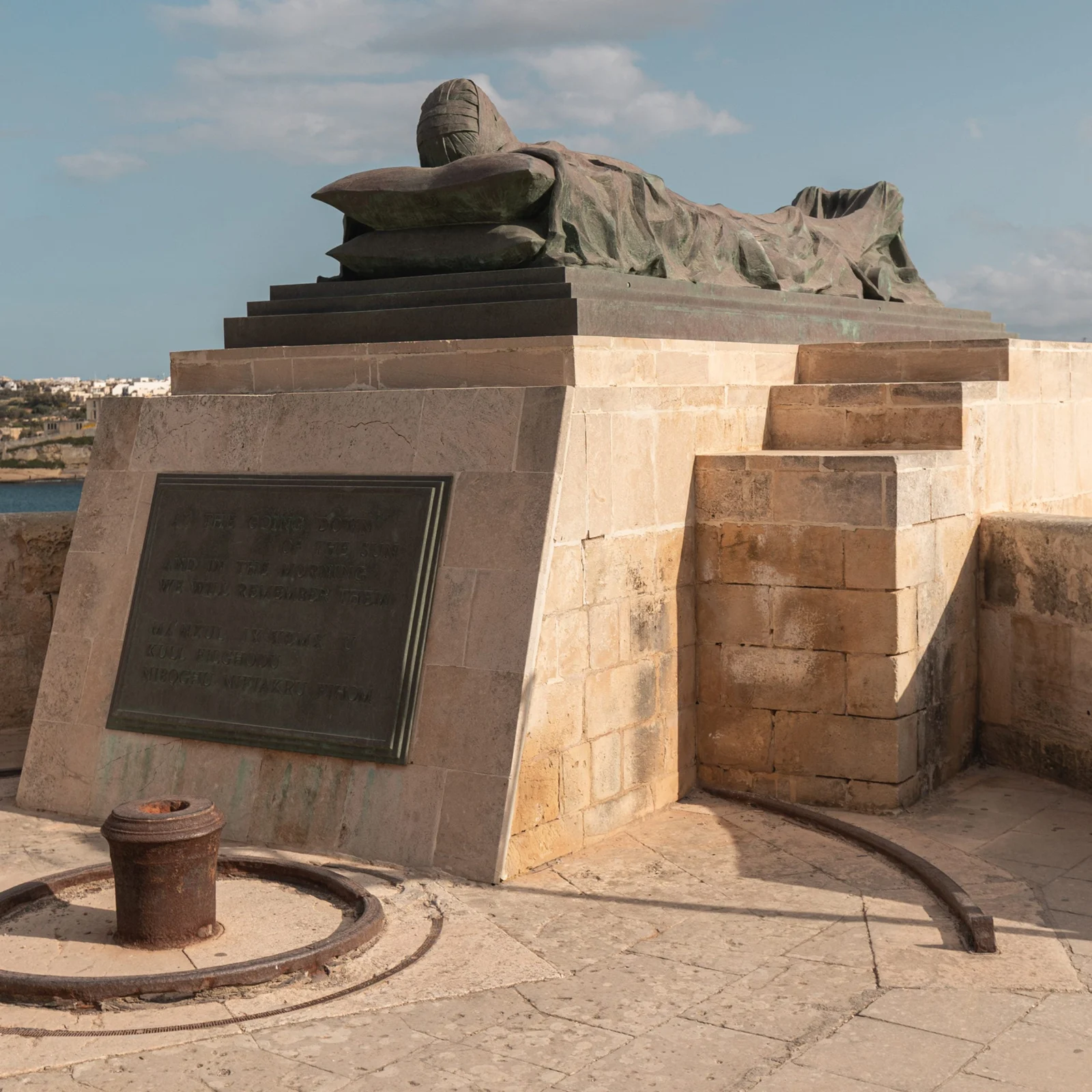 A bronze figure lies atop a stone monument bearing a memorial plaque, overlooking the sea under a partly cloudy sky.