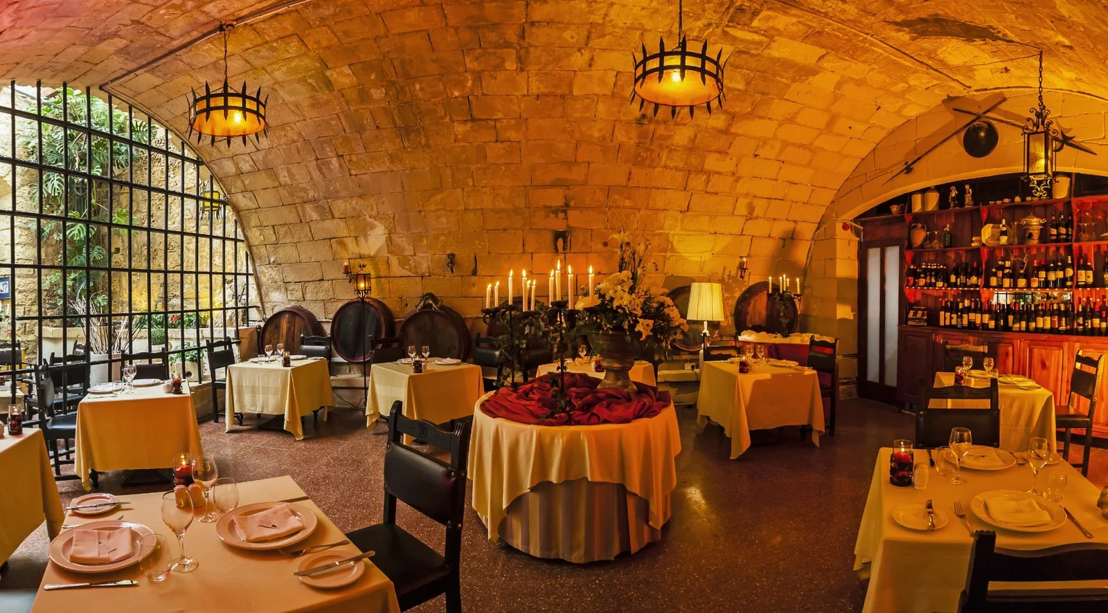 A candlelit stone-vaulted restaurant dining room with set tables, wine barrels and a well-stocked bar along the back wall.