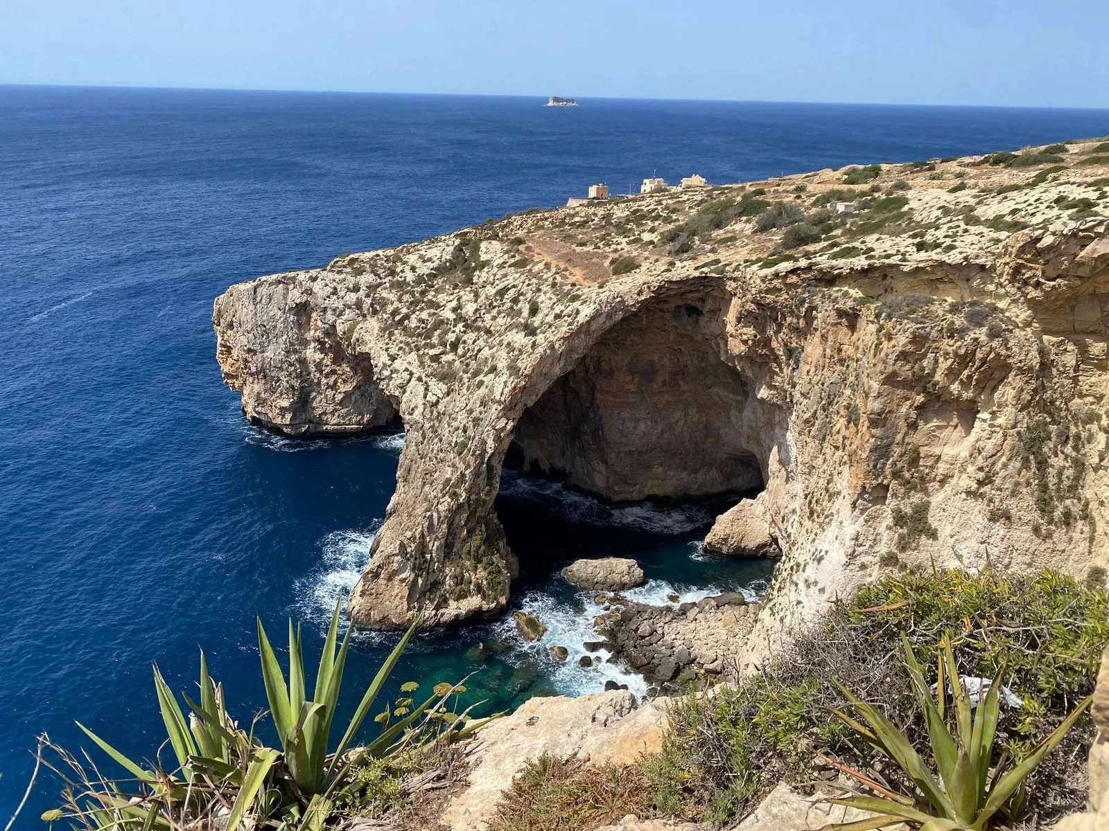 A large natural rock arch over vivid blue sea beside limestone cliffs with sparse vegetation under a clear sky.