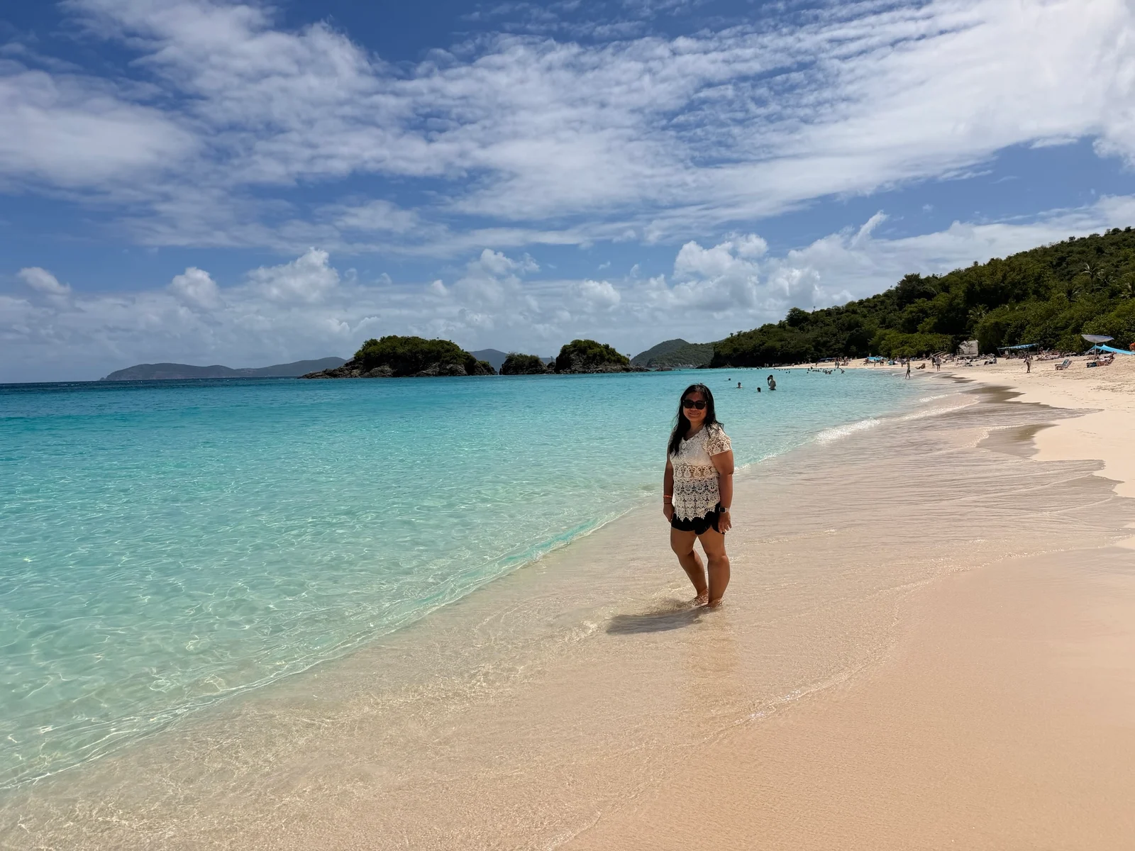 A woman in a white lace top and black shorts stands at the edge of clear turquoise water on a sandy tropical beach.