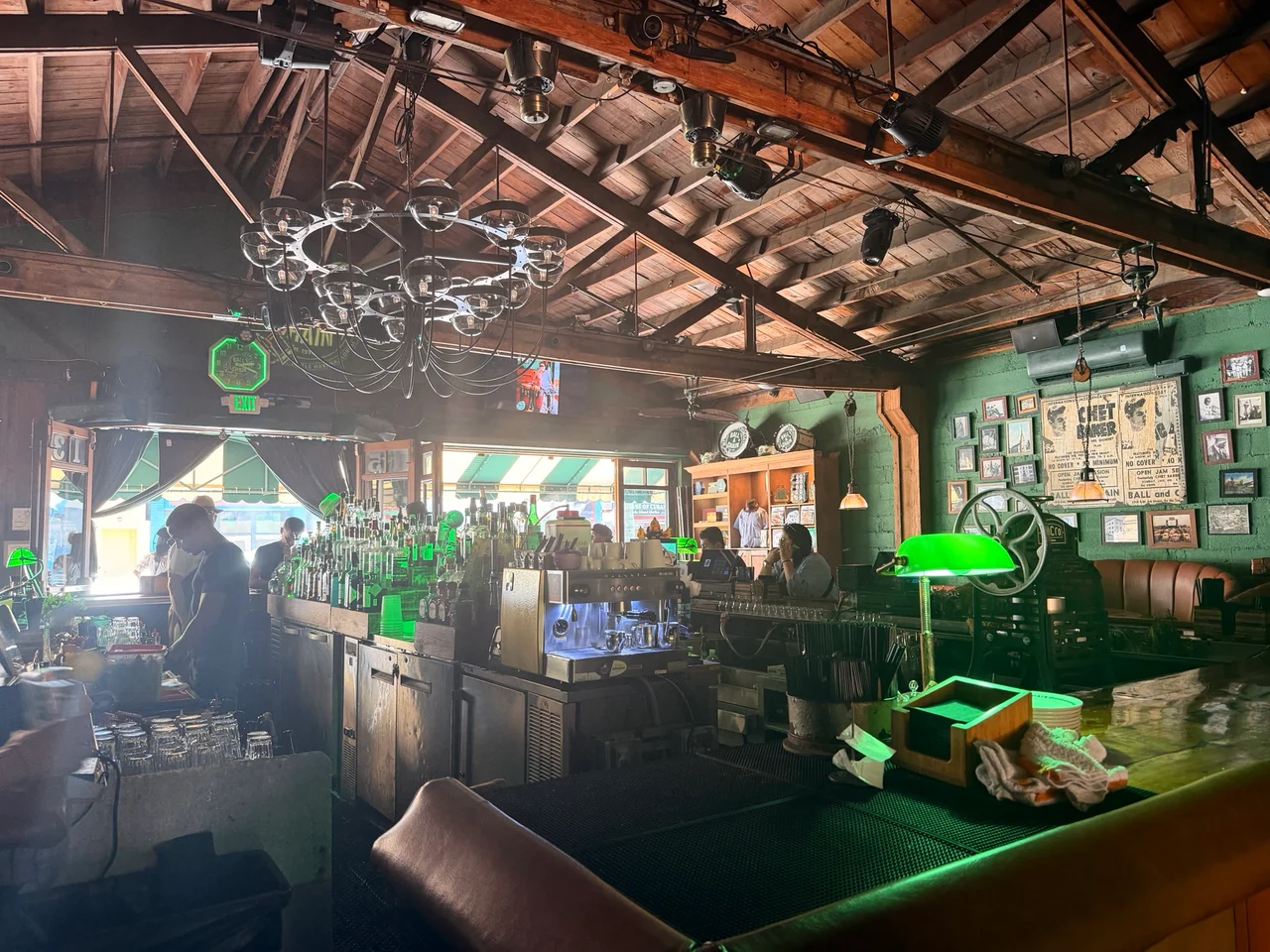 A dimly lit bar interior with wooden ceiling beams, green walls, green bankers lamps and bottles lined along the counter.