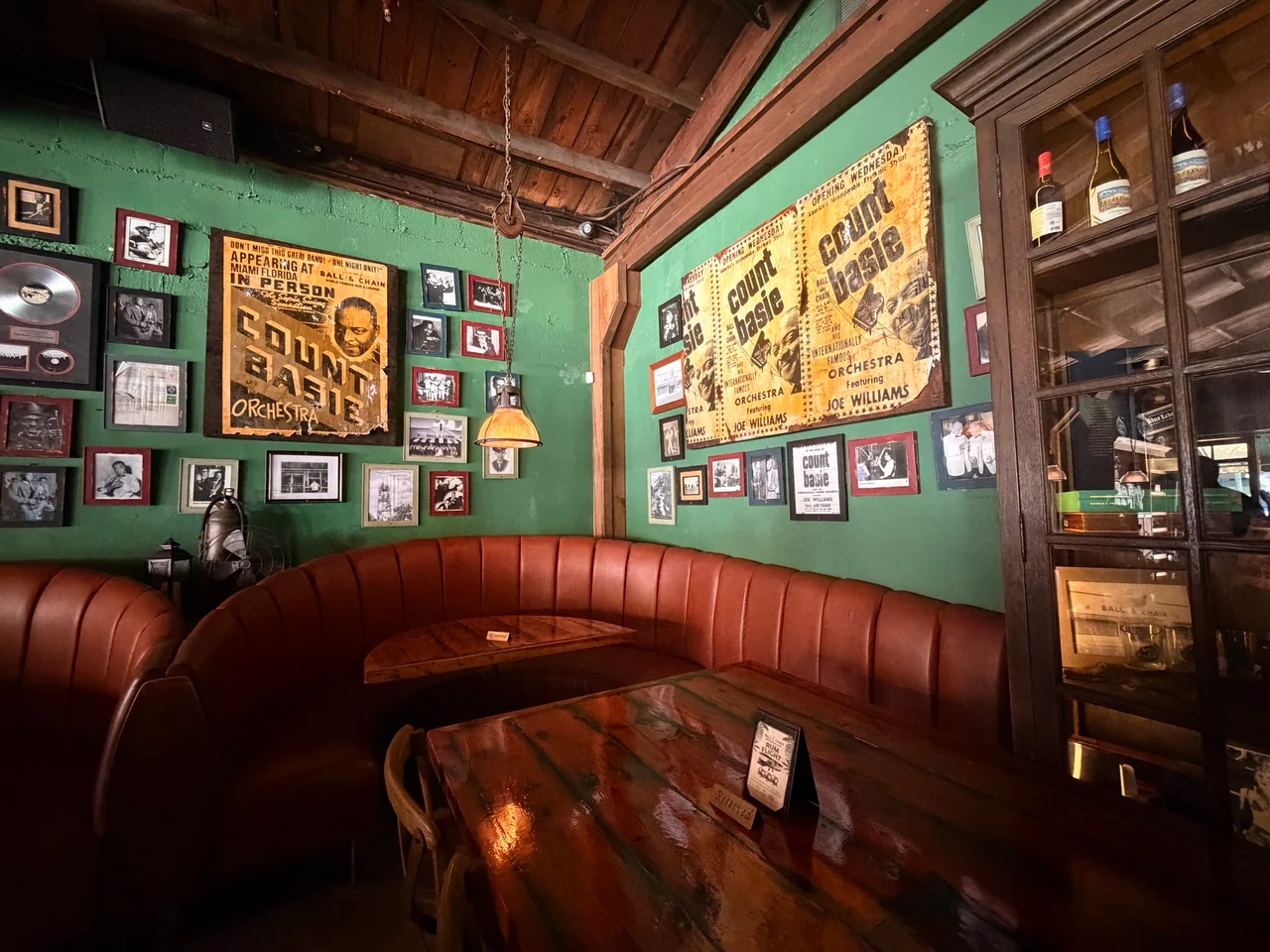 A curved red leather booth beneath vintage Count Basie concert posters and framed photographs on green walls in a dimly lit bar.