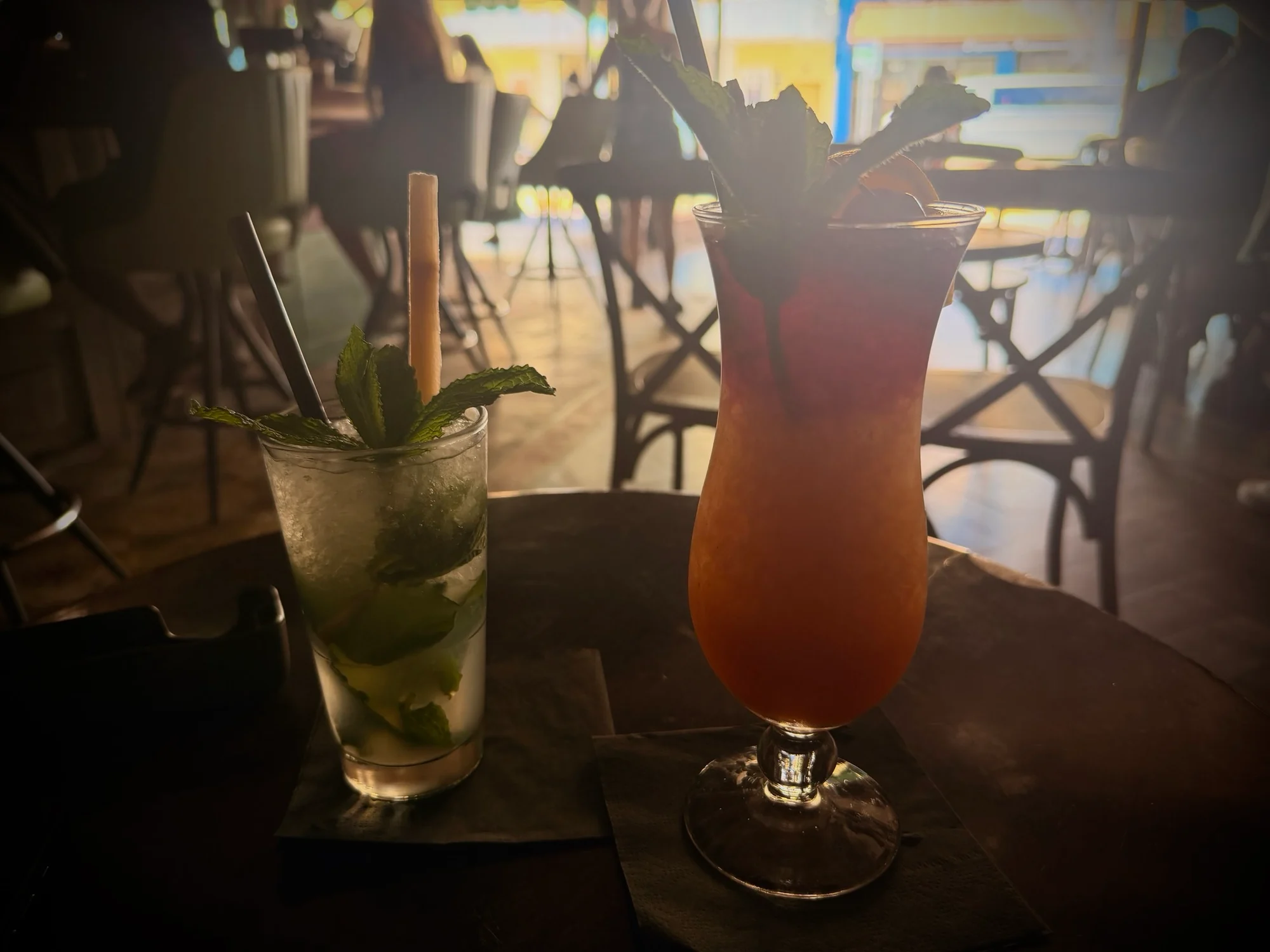 Two cocktails on a dark wooden table, a mojito with mint leaves and a tall red drink with pineapple garnish.