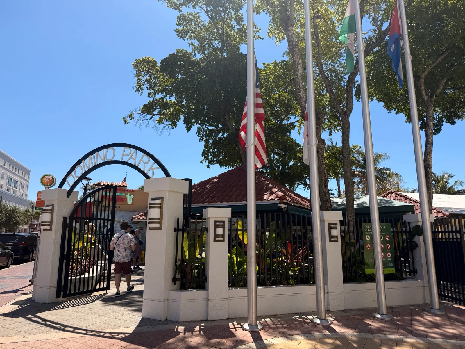 The entrance gate to Domino Park with an arched sign, black iron railings and three flags on poles under a clear blue sky.