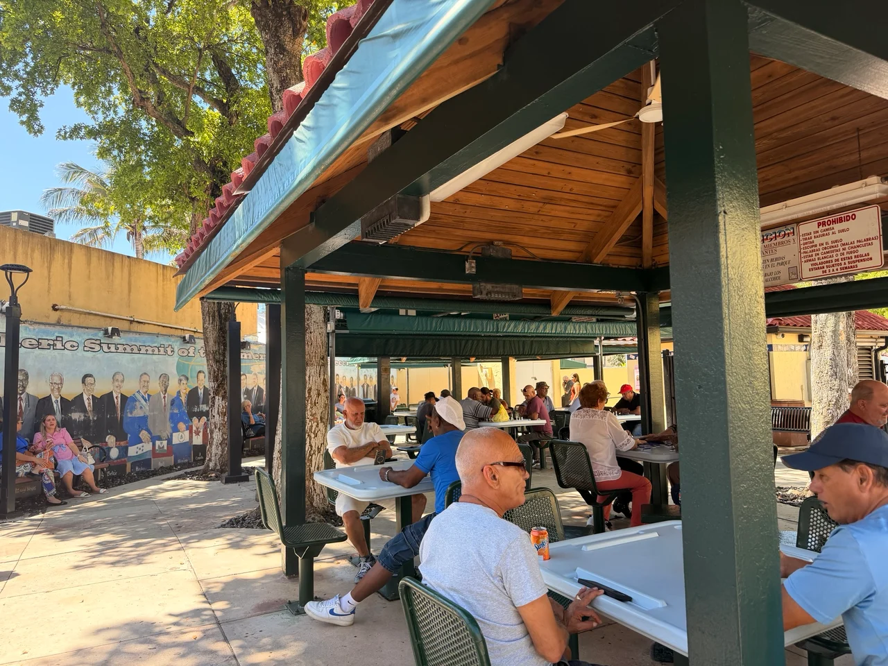 People sit beneath a covered outdoor pavilion at tables, with a colourful mural visible on a wall behind them.