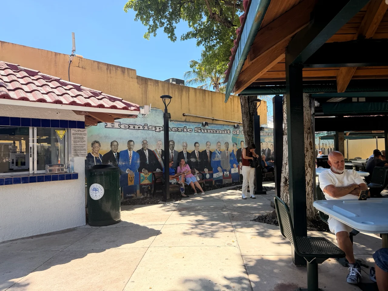 Outdoor dining area with a mural reading Hemispheric Summit on a yellow wall, beside a small service kiosk.
