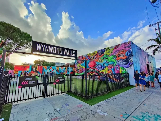 The entrance to Wynwood Walls with a black metal fence and a colourful graffiti mural on the building behind it.