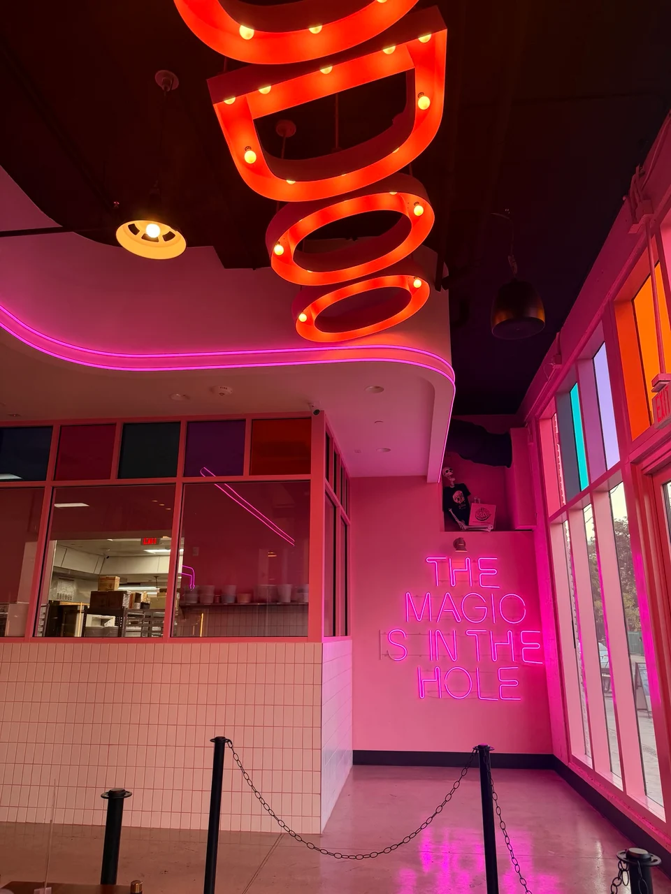 Interior of a pink neon-lit doughnut shop with a glowing sign reading The Magic Is In The Hole on the wall.