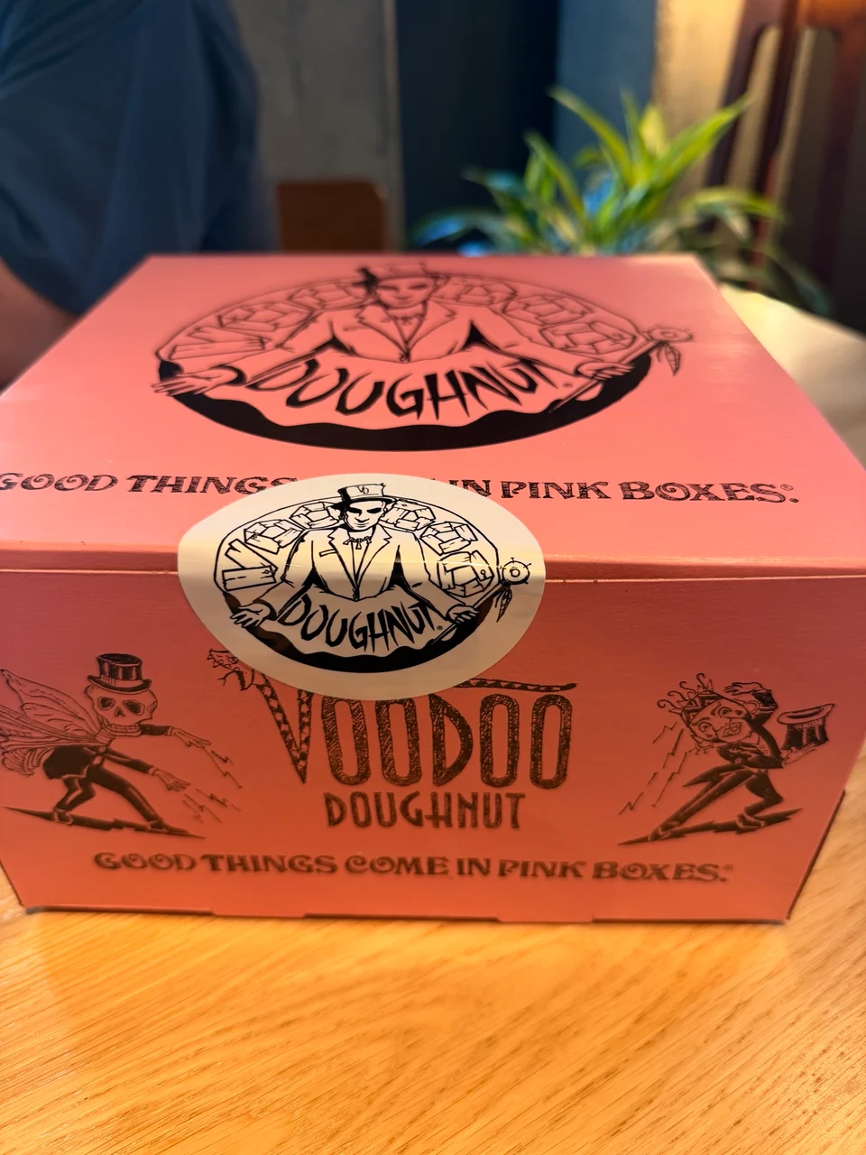 A pink Voodoo Doughnut box on a wooden table with the slogan Good Things Come in Pink Boxes printed on the side.