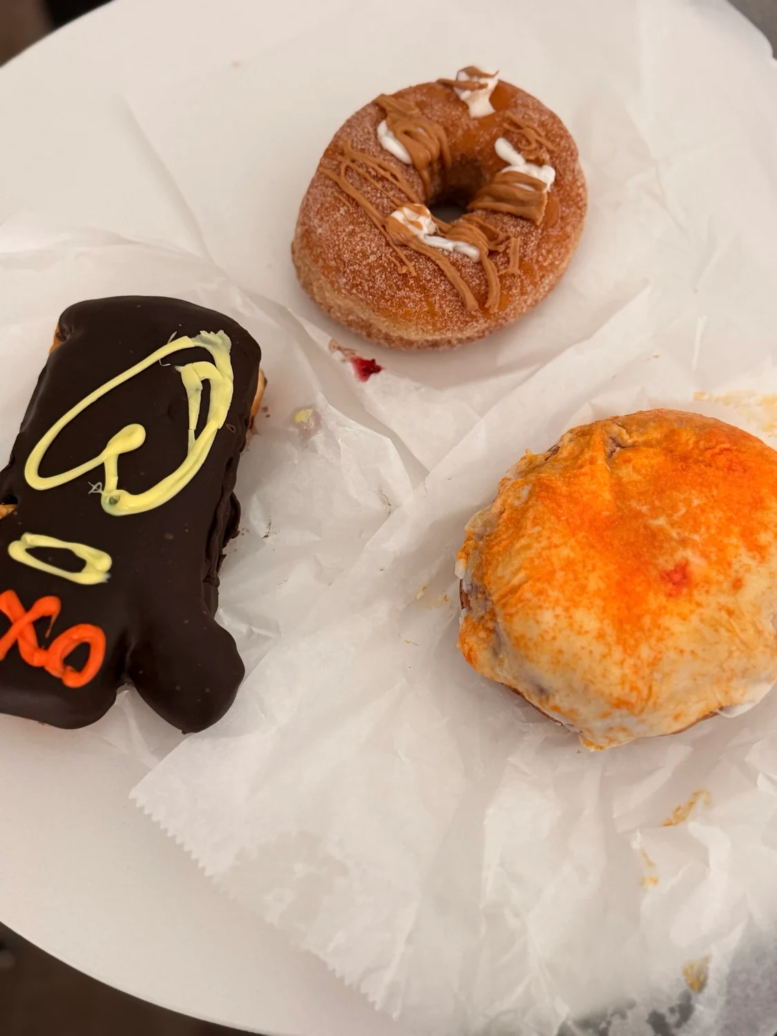 Three pastries on waxed paper: a sugared ring doughnut, a dark chocolate bear-shaped treat and a golden cheesy scone.