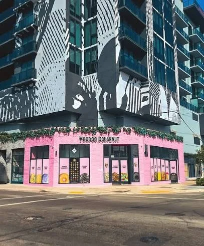 A pink corner building with a Voodoo Doughnut sign sits beside a tall apartment block covered in a large black and white mural.