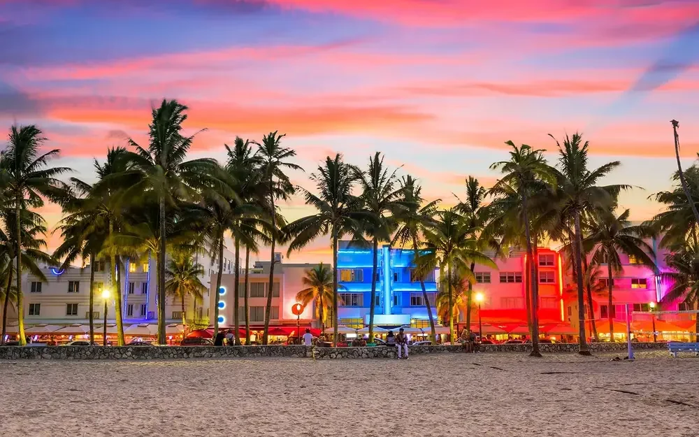 A sandy beach at sunset lined with palm trees and neon-lit Art Deco buildings glowing in pink, blue and orange.