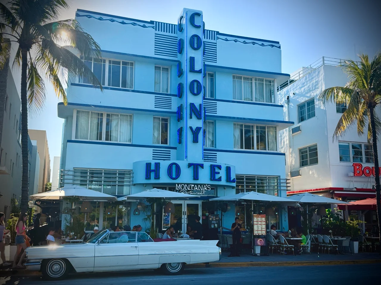 A white Art Deco Colony Hotel building with a blue neon sign, outdoor dining and a white classic convertible parked outside.