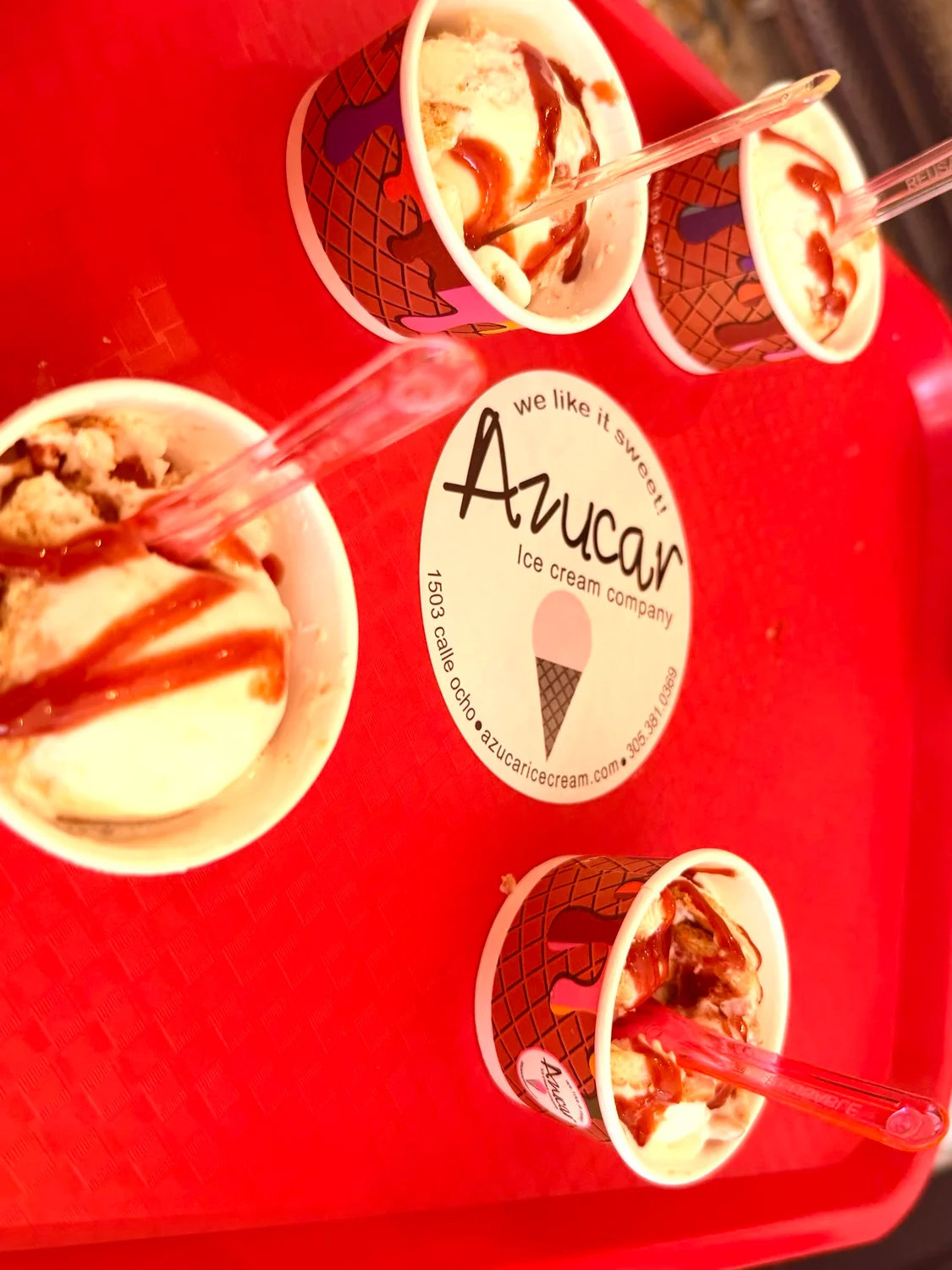 Four cups of ice cream with syrup and plastic spoons on a red tray bearing an Azucar Ice Cream Company sticker.