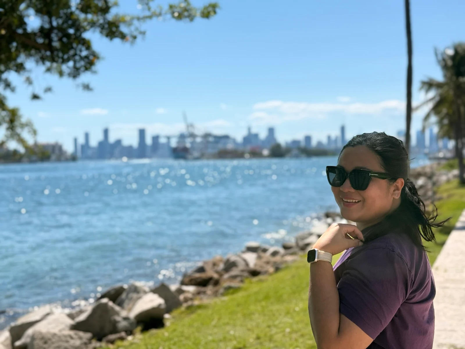 A woman in sunglasses and a purple top smiles over her shoulder beside a rocky waterfront with a city skyline behind her.