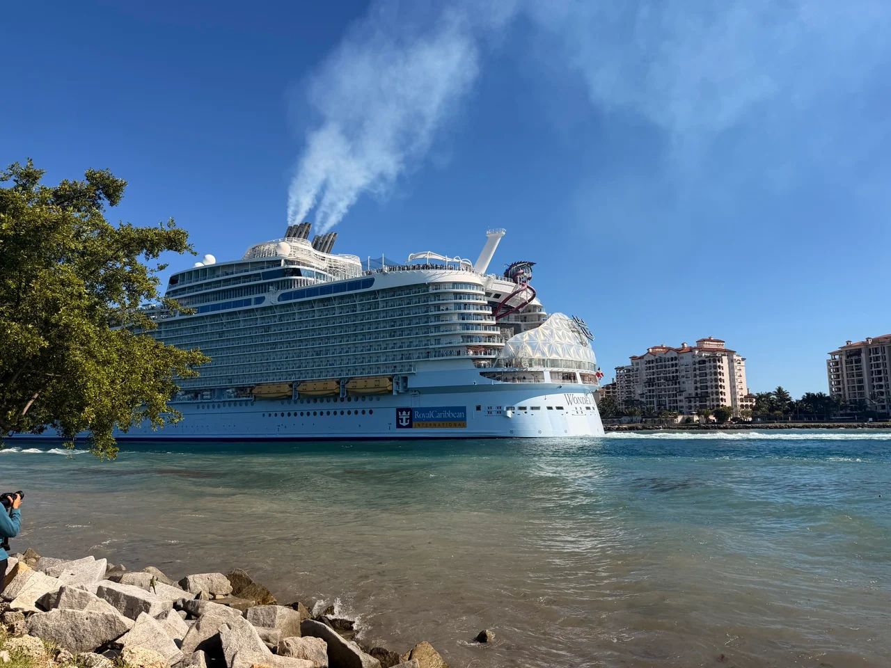 A Royal Caribbean cruise ship named Wonder sails past a rocky shoreline under a clear blue sky, with smoke rising from its funnels.