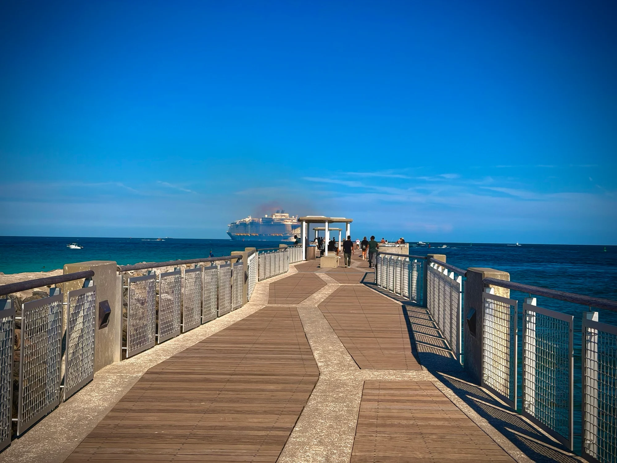 A wooden pier stretches toward the sea where a large cruise ship sails under a clear blue sky.