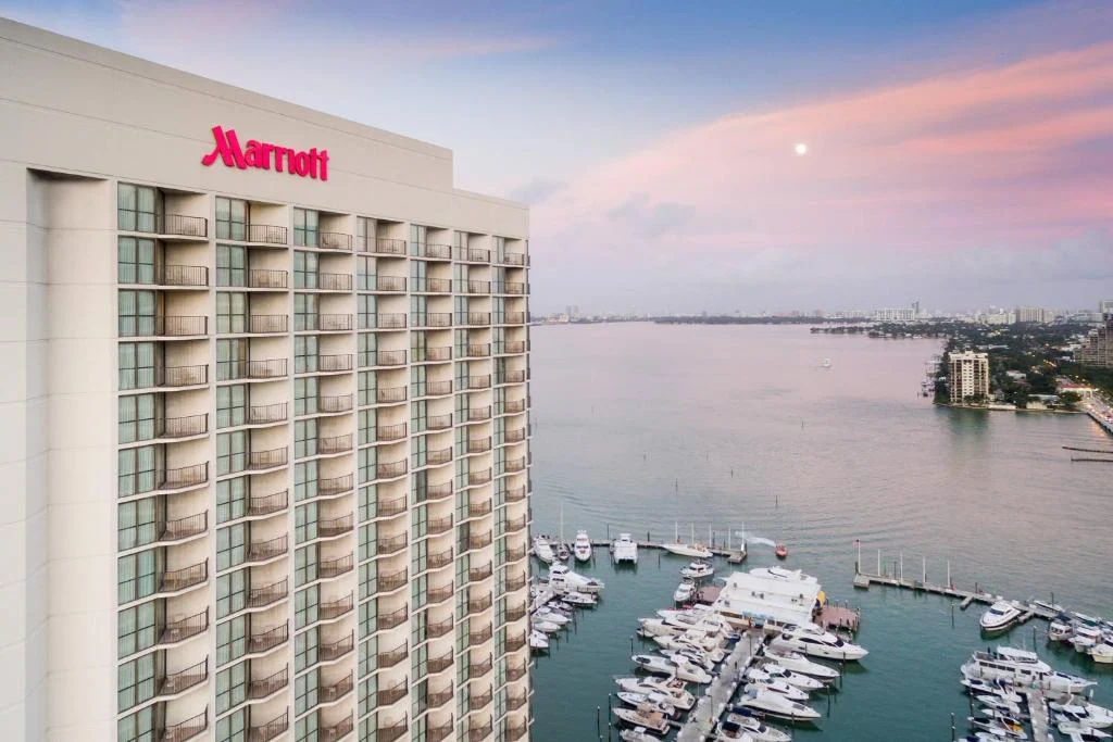 A tall Marriott hotel with balconies overlooks a busy marina and a calm bay at dusk under a pink sky.