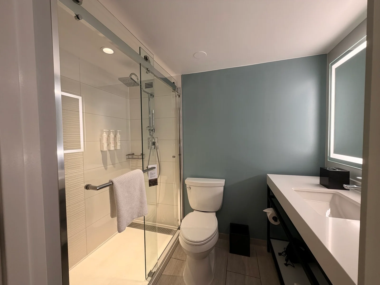 A modern hotel bathroom with a glass walk-in shower, white toilet, long vanity with white countertop and a lit mirror.