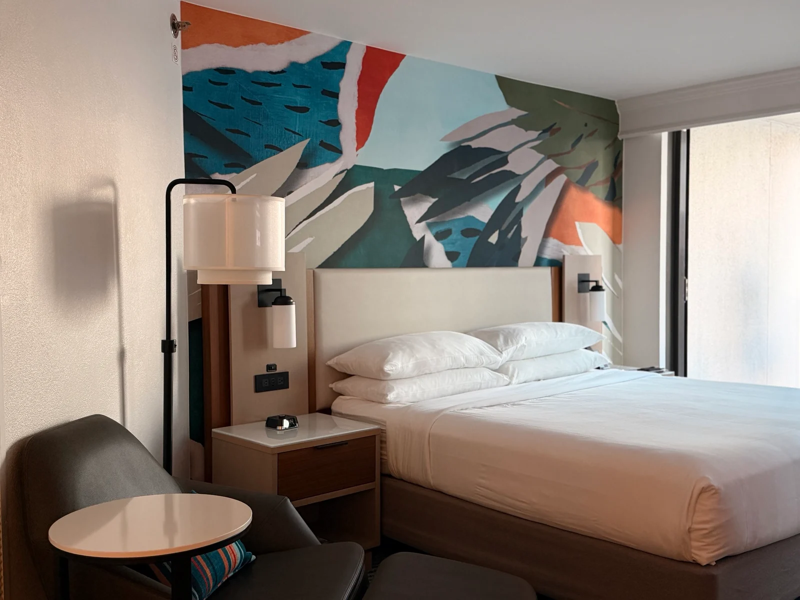 A hotel-style room with a king bed, white bedding, a floor lamp and a colourful abstract mural on the wall behind the headboard.