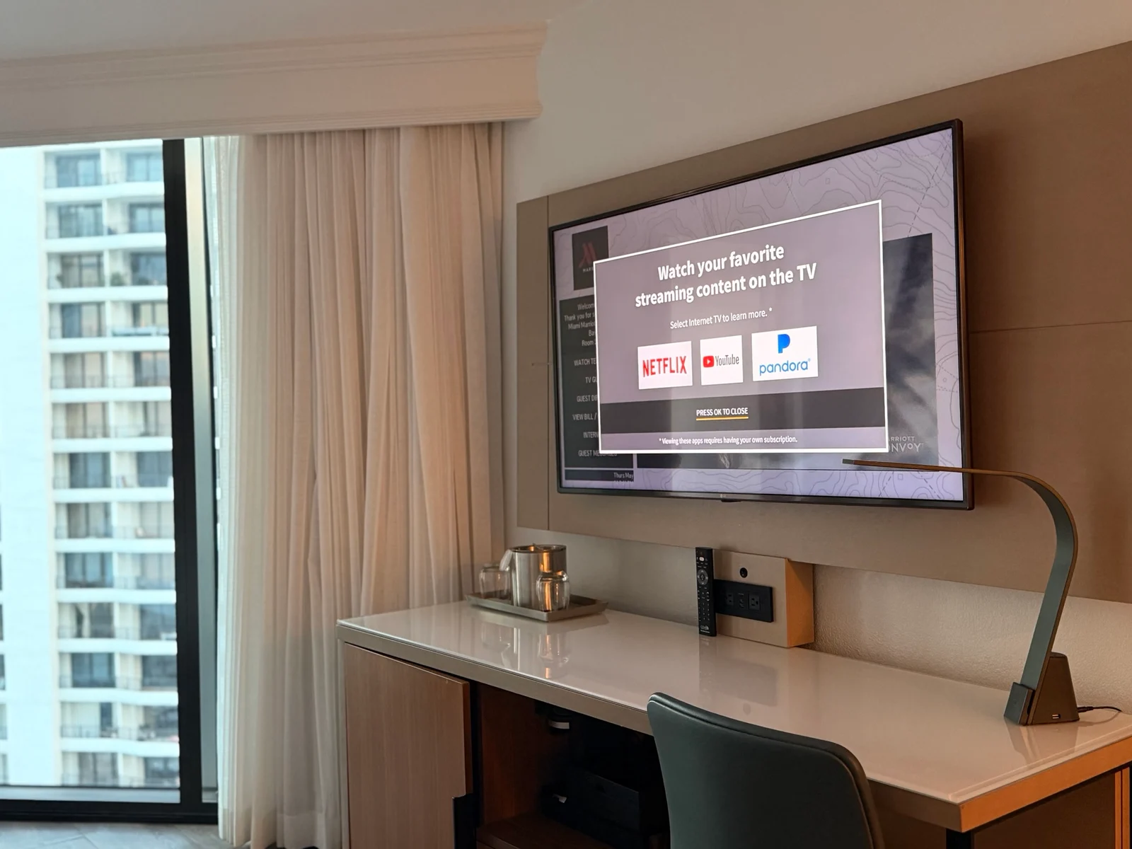 A hotel room TV on a wall shows a streaming services welcome screen above a white desk with a remote control and lamp.