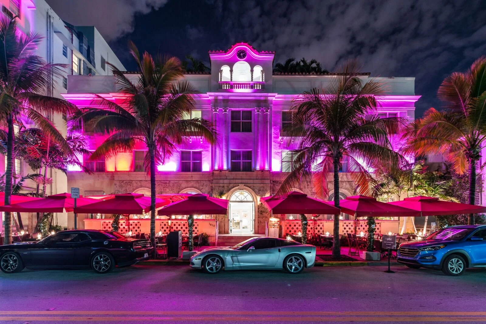 A white Art Deco hotel lit with vivid pink and purple lights at night, with palm trees and red parasols outside.