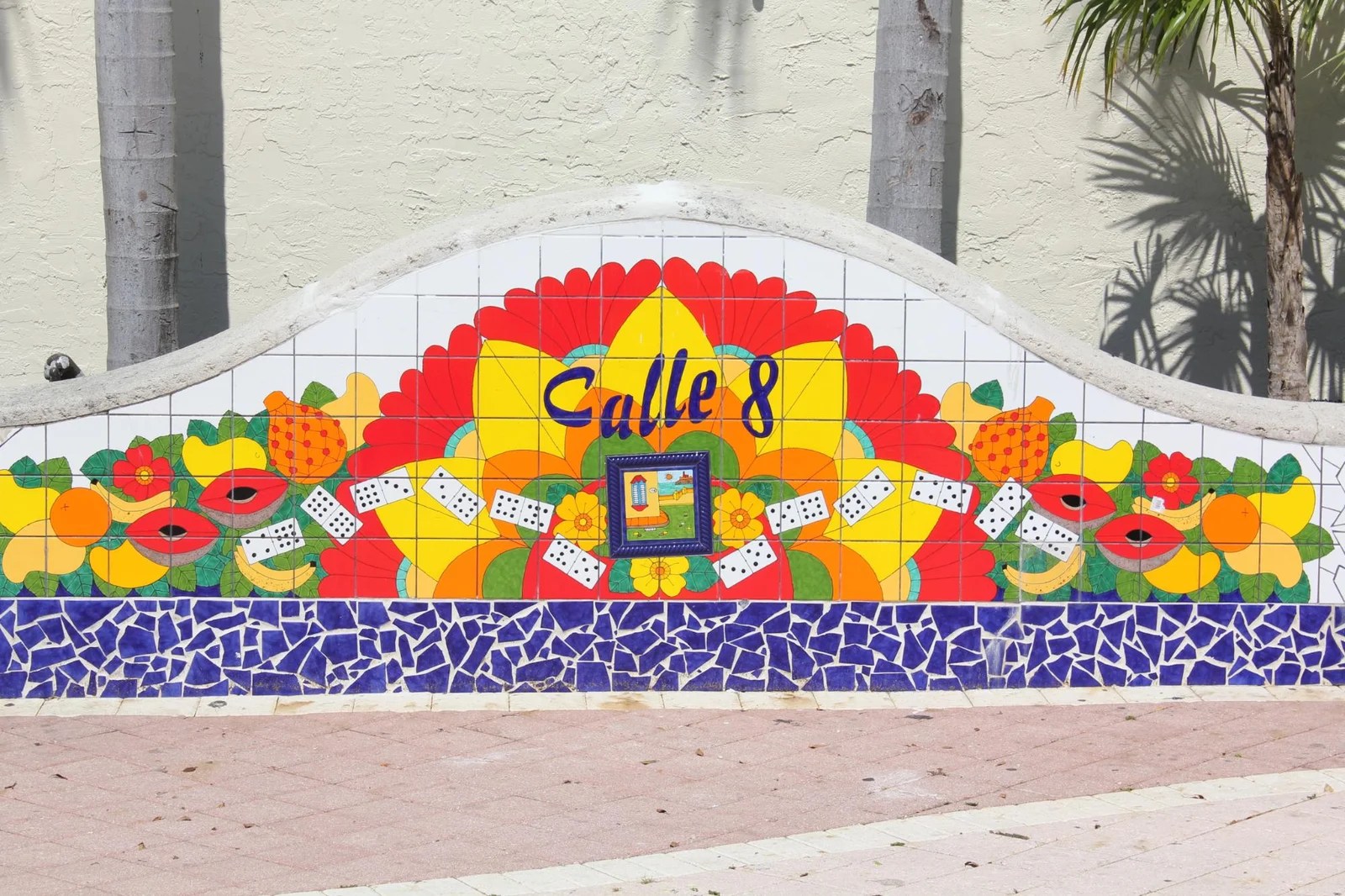 A colourful tiled mosaic wall reading Calle 8, decorated with fruit, dominoes and flowers, with palm trees behind.