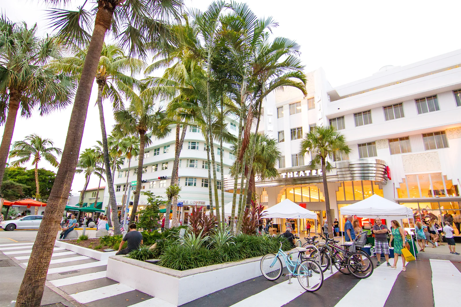 Palm-lined street with parked bicycles, white gazebos and a theatre entrance flanked by Art Deco buildings and pedestrians.
