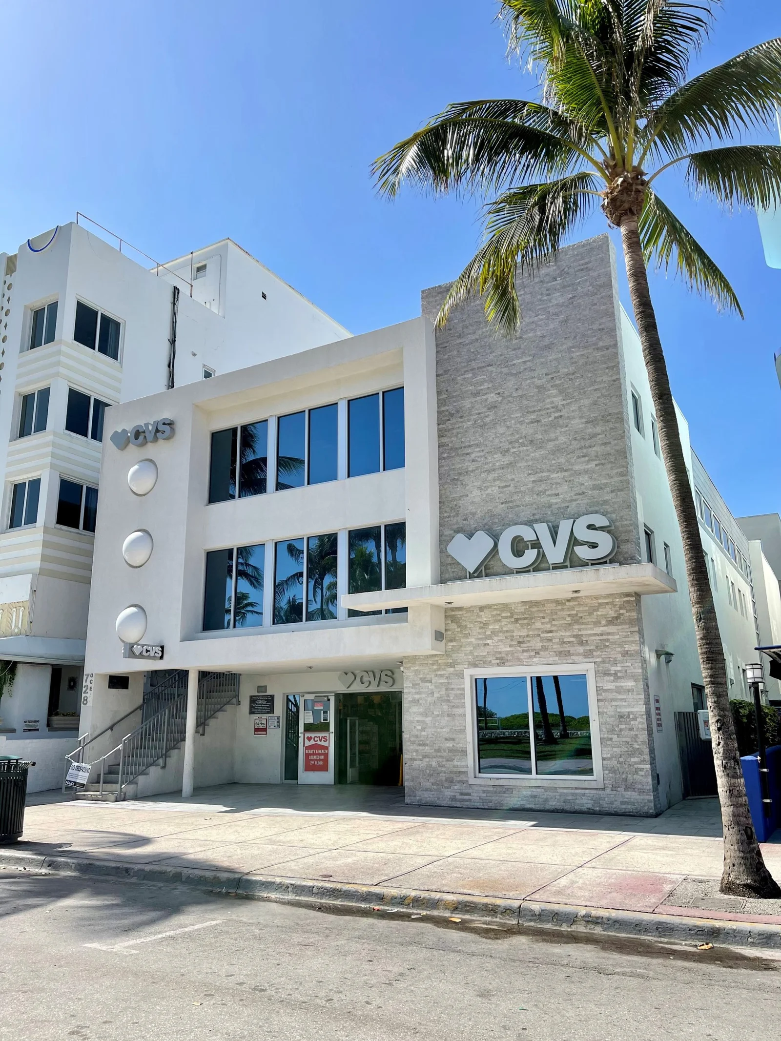 A white multi-storey CVS pharmacy building with large windows and a tall palm tree on a sunny day.