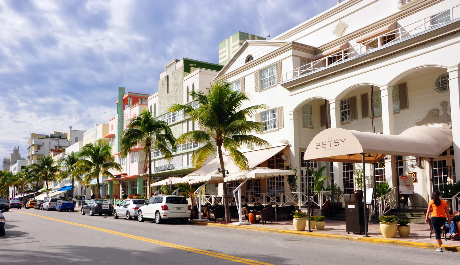 A sunny street lined with palm trees, Art Deco hotels and parked cars, with a woman walking past the Betsy hotel.