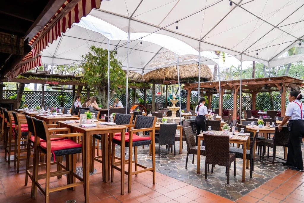 An outdoor restaurant patio with wooden tables, red-cushioned chairs and a thatched canopy covered by a white marquee tent.