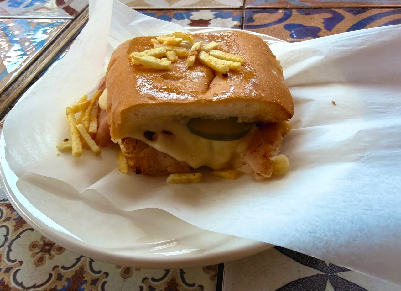 A chicken burger with melted cheese, pickles and chips stuffed inside a glazed bun, served on a white plate lined with paper.
