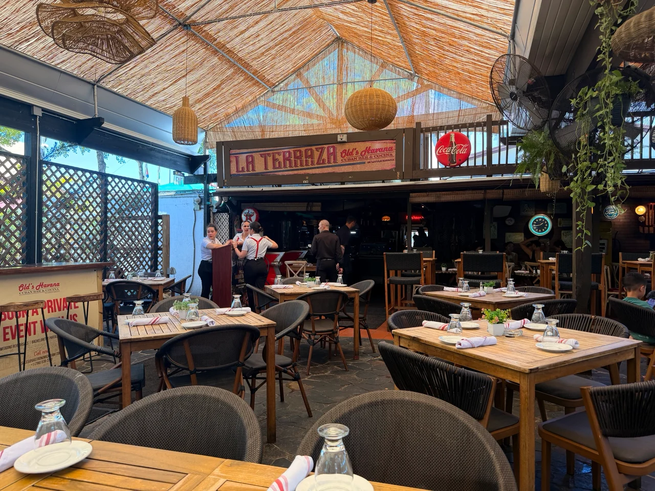 La Terraza Cuban Bar and Cocina restaurant with wicker ceiling, wooden tables and staff in red braces standing near the bar.