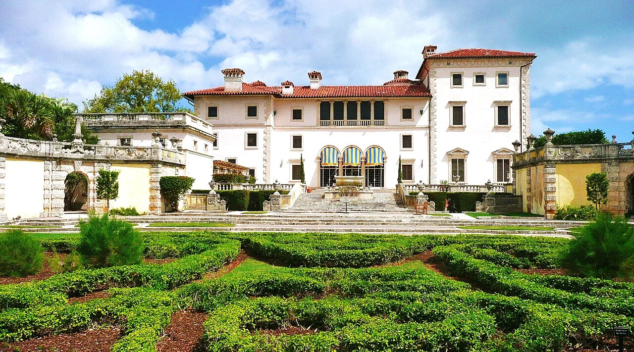 A grand white Italian-style villa with a red-tiled roof, stone staircases and formal box-hedge gardens in the foreground.