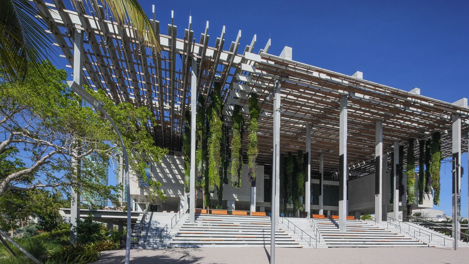 A modern building with a latticed canopy and hanging plants above wide steps, surrounded by trees under a clear blue sky.